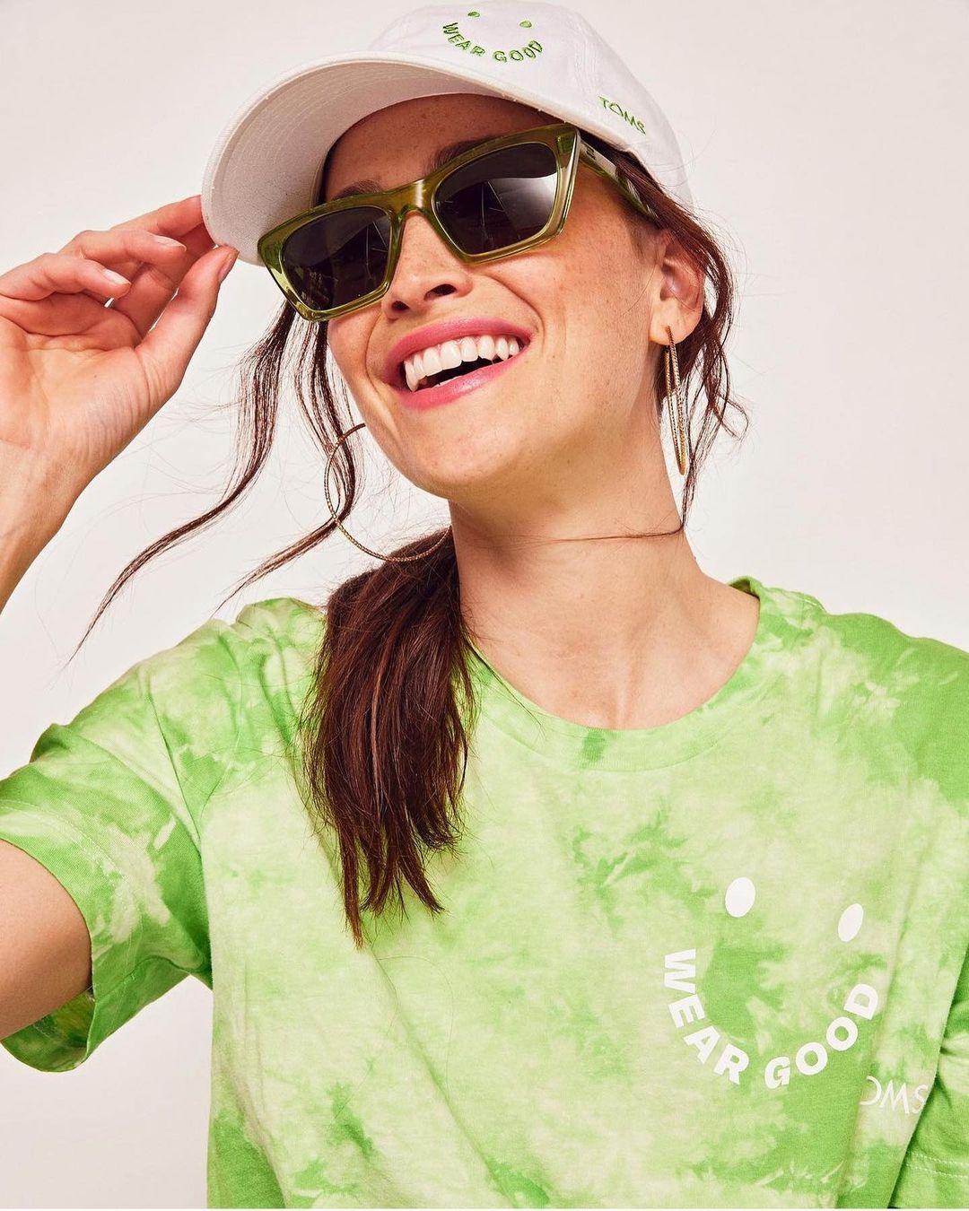 Smiling woman in tie-dye t-shirt and white baseball cap, wearing olive green sunglasses, posing against a neutral backdrop.