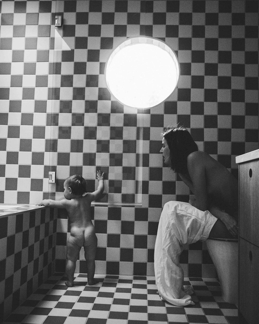 Black and white photo in a tiled bathroom of a mother sitting on the toilet and a baby standing by the bathtub.