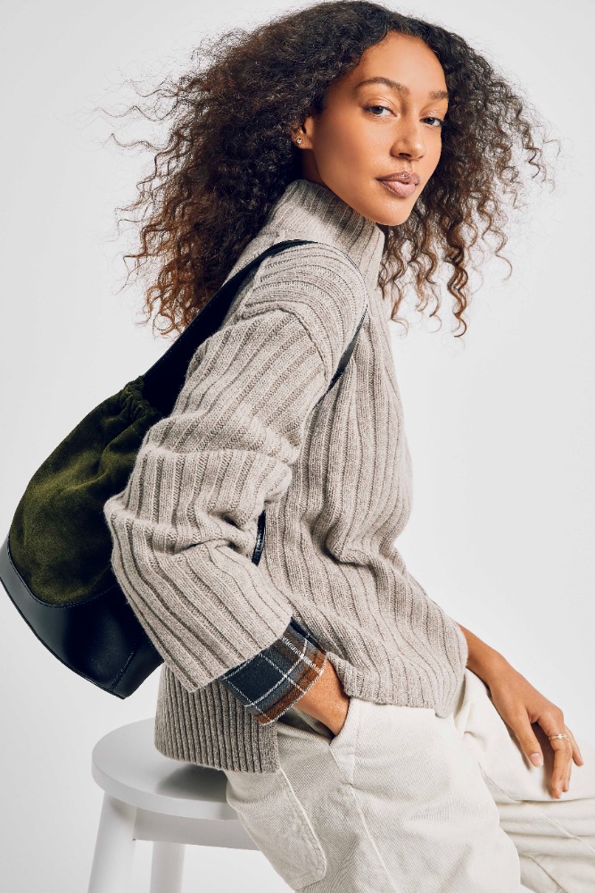 Attractive brunette woman poses indoors wearing ribbed sweater, beige pants, and black tote bag against a neutral backdrop.