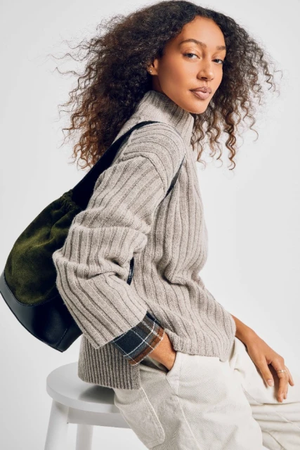 Attractive brunette woman poses indoors wearing ribbed sweater, beige pants, and black tote bag against a neutral backdrop.