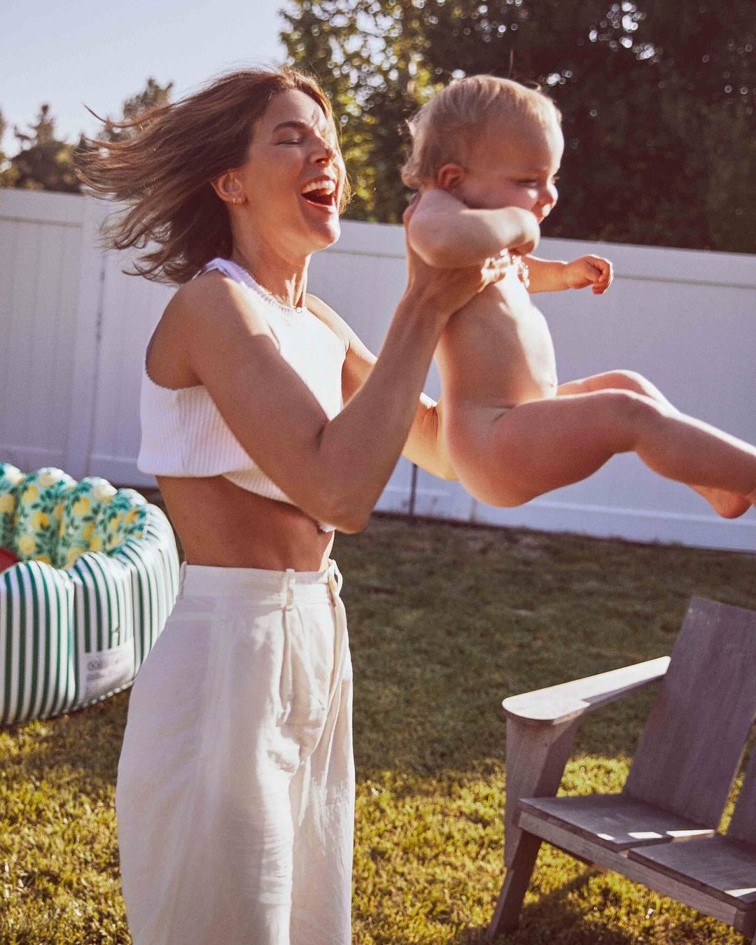 Woman lifts a baby high into the air outdoors on grass in bright sunlight near a white fence. A small pool sits in the background.