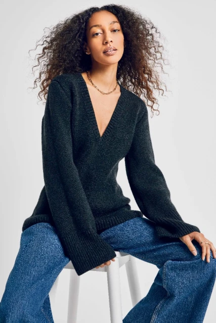 Portrait of a woman with curly hair wearing a gray sweater and blue jeans, seated on a white stool against a white backdrop.
