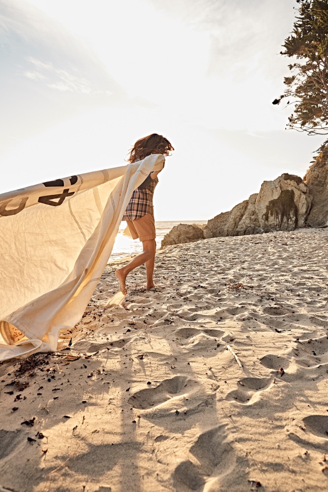 Woman running barefoot on sandy beach holding large piece of fabric catching wind