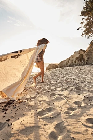 Woman running barefoot on sandy beach holding large piece of fabric catching wind