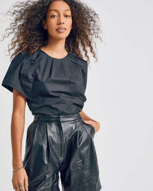 Brunette woman in a black blouse and black leather shorts against a plain white background in a studio shot. Fashionable look.