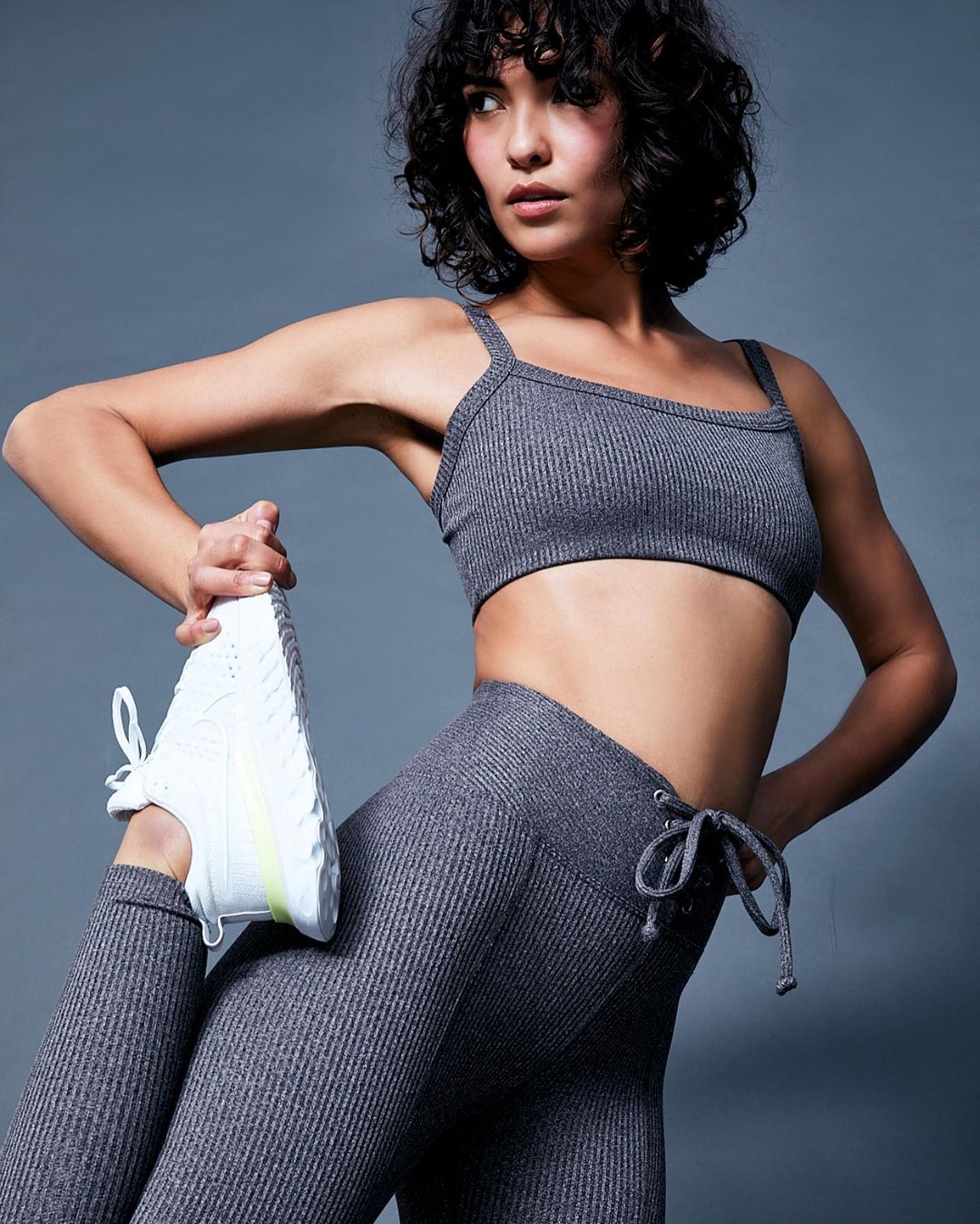 Athletic woman in grey ribbed fitness set stretches leg holding white sneaker against grey backdrop in medium studio shot.