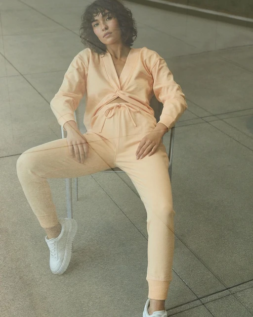 A young woman with curly dark hair poses in a peach-colored lounge set and white sneakers while sitting in a metal chair against a concrete wall.