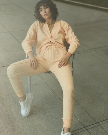 A young woman with curly dark hair poses in a peach-colored lounge set and white sneakers while sitting in a metal chair against a concrete wall.