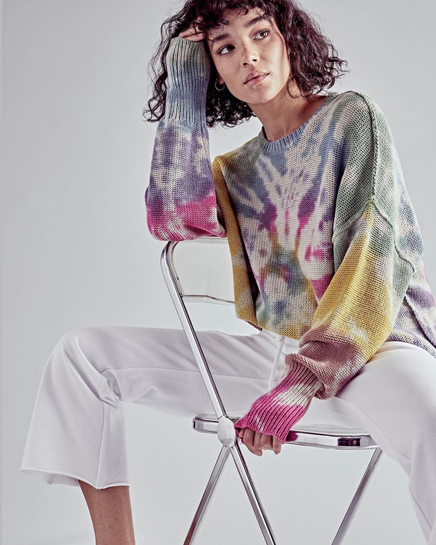Woman seated, wearing colorful tie-dye sweater and white pants in a studio, posing in front of white backdrop with soft, diffused lighting.
