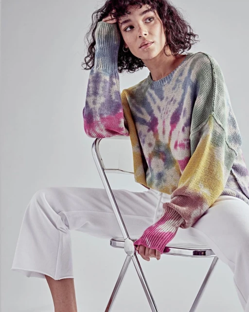 Woman seated, wearing colorful tie-dye sweater and white pants in a studio, posing in front of white backdrop with soft, diffused lighting.