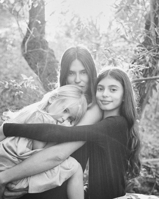Black and white portrait of a mother holding her two daughters in a natural outdoor setting with dappled light filtering through trees.