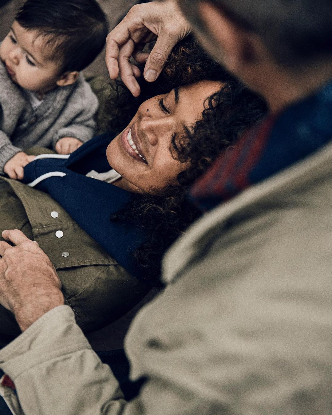 A smiling woman with dark curly hair reclines with her eyes closed while a baby and older man look at her affectionately.