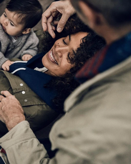 A smiling woman with dark curly hair reclines with her eyes closed while a baby and older man look at her affectionately.