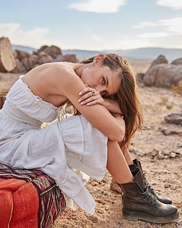 Woman in white ruffled dress and black combat boots seated outdoors in a desert landscape by Annie Edmonds