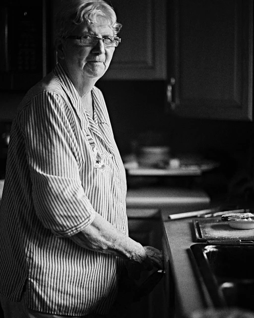 Older woman in a striped shirt and glasses stands inside a kitchen next to a sink in a black and white medium shot.
