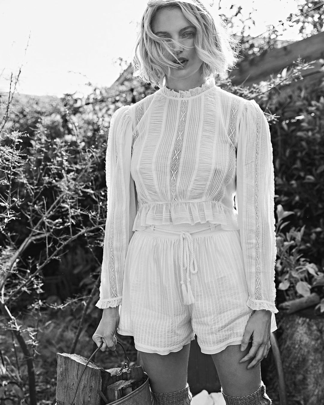 Monochrome portrait of a woman in a white lace top and shorts in an outdoor setting with natural light, holding a metal bucket.