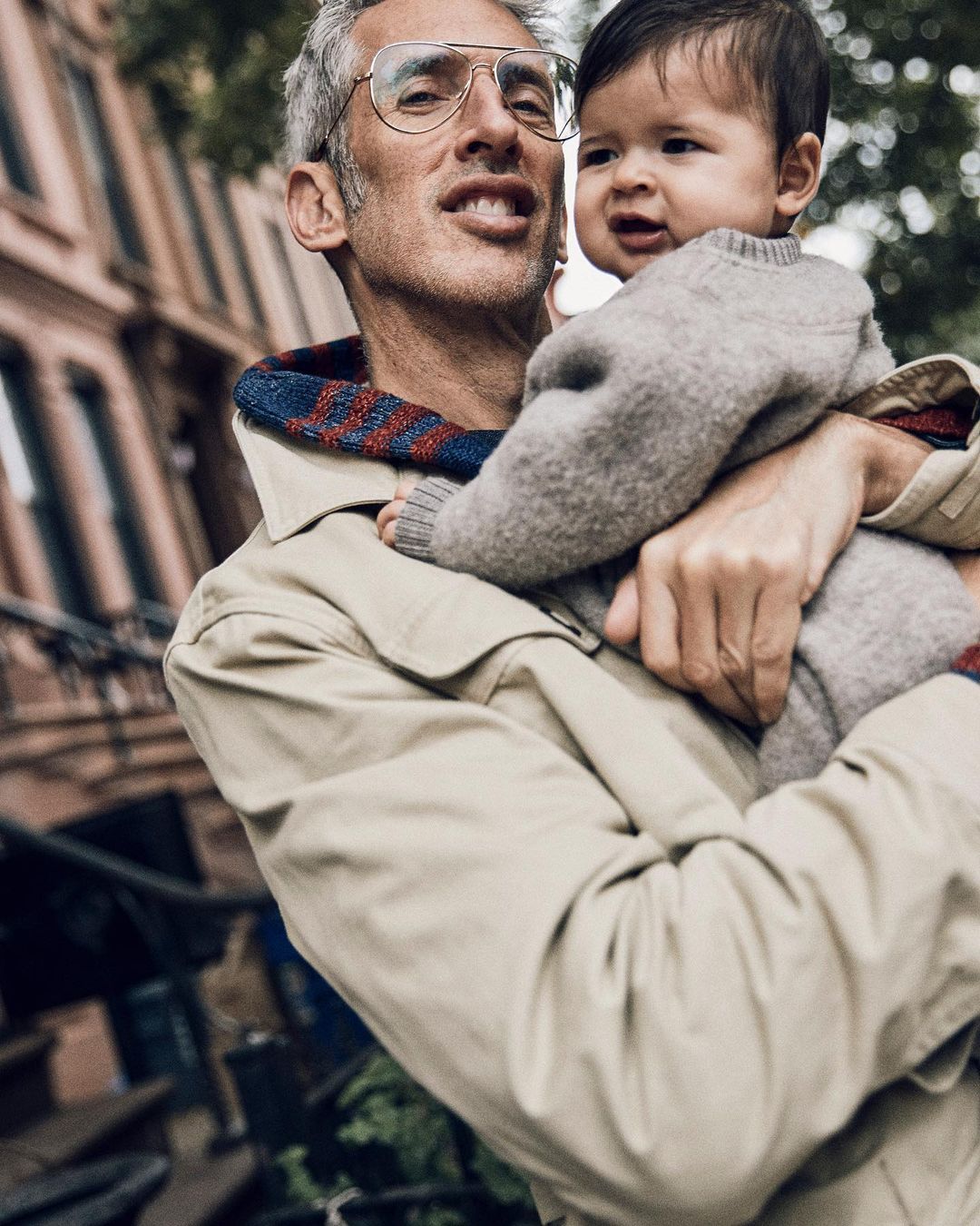 Adult male in eyeglasses holding a baby wearing a sweater outdoors in front of building in daylight. Father and child portrait.