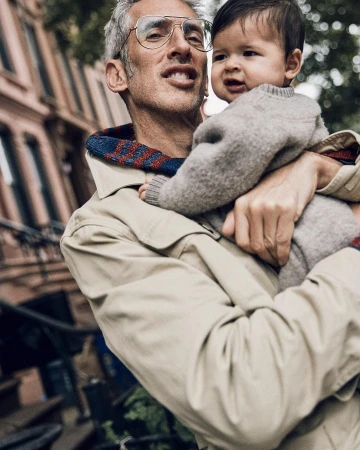 Adult male in eyeglasses holding a baby wearing a sweater outdoors in front of building in daylight. Father and child portrait.