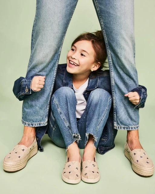 A smiling girl in a jean jacket sits cross-legged between the legs of a person wearing jeans and shoes with a light green background.