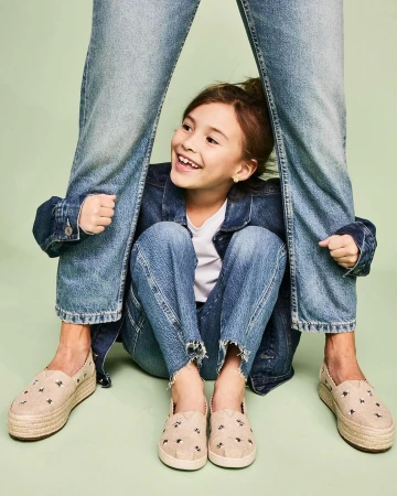 A smiling girl in a jean jacket sits cross-legged between the legs of a person wearing jeans and shoes with a light green background.