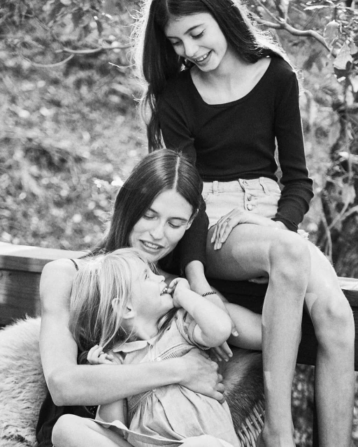 Black-and-white shot of a mother embracing her young daughter while her older daughter stands beside them, all smiling in an outdoor setting.