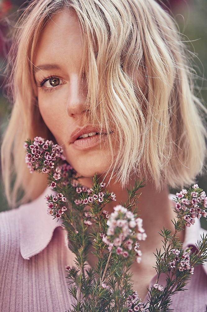 Portrait of a blonde woman with bob haircut holding a flowering plant with small pink blossoms against a blurred, natural background.