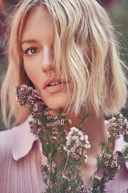 Portrait of a blonde woman with bob haircut holding a flowering plant with small pink blossoms against a blurred, natural background.