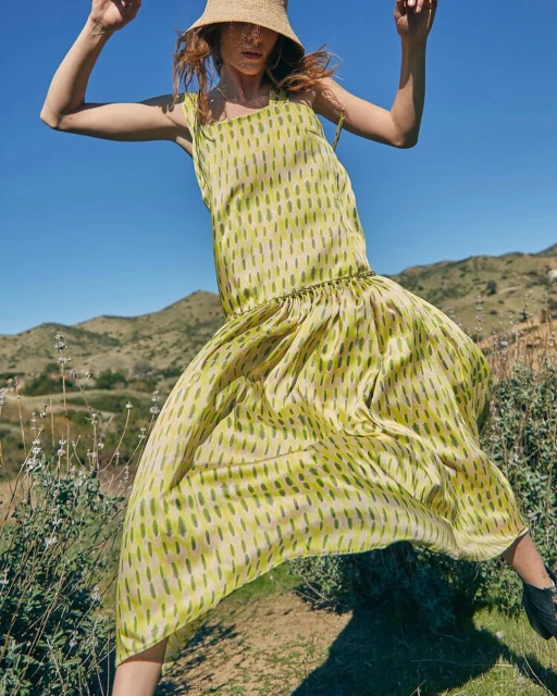 A woman in a yellow patterned dress and straw hat jumps happily in a sunny, hilly landscape. The style evokes a sense of summer freedom.