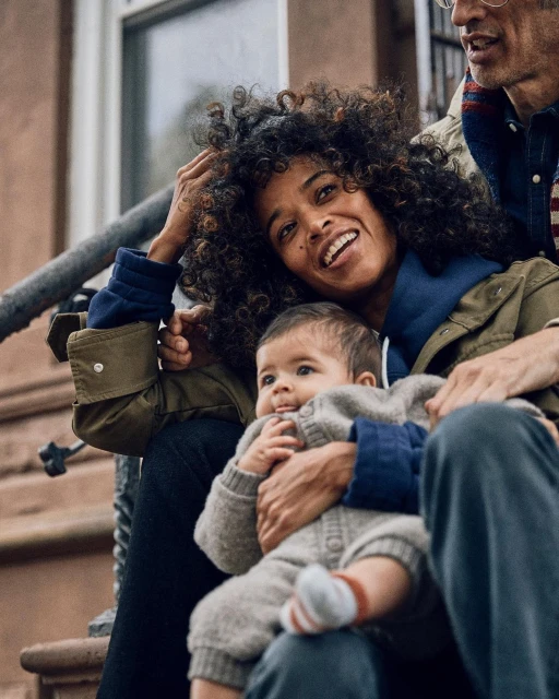 A mother and child, sitting closely on outdoor steps in daytime. The mother has dark curly hair, and the baby is wearing a grey onesie.