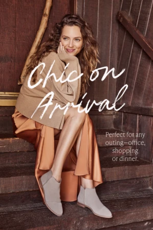 Woman with long curly brown hair seated on wooden steps wearing a beige sweater, satin skirt, and grey ankle boots.