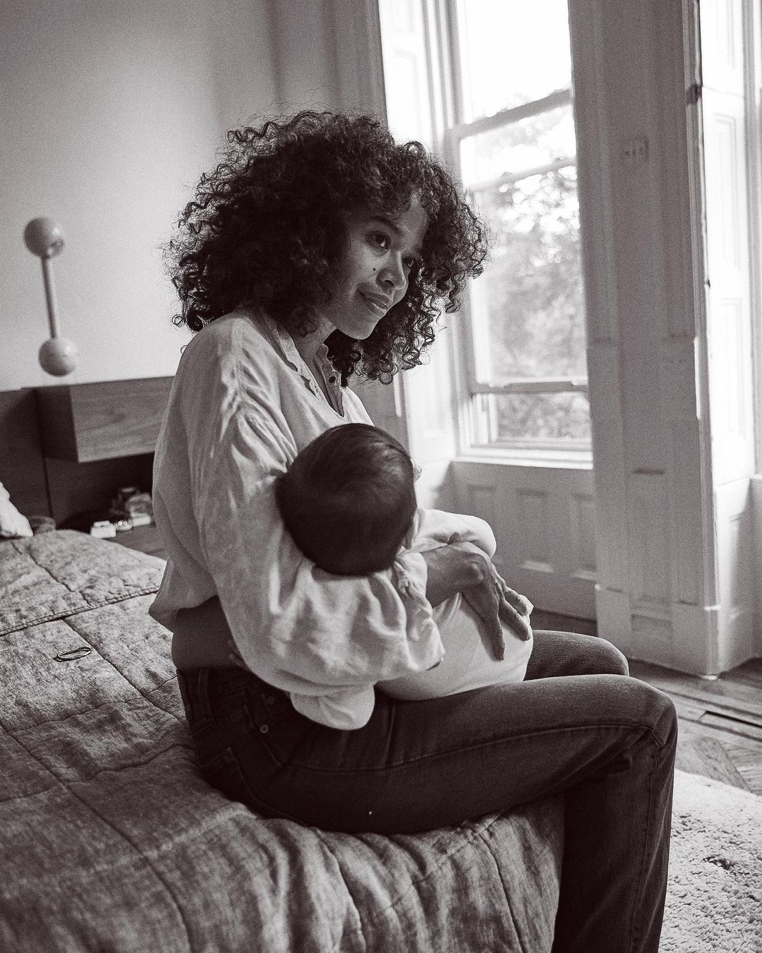Monochrome portrait of an adult female sitting on a bed, holding a baby in her arms near a window providing natural light.