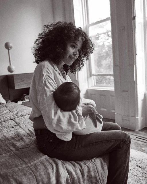 Monochrome portrait of an adult female sitting on a bed, holding a baby in her arms near a window providing natural light.