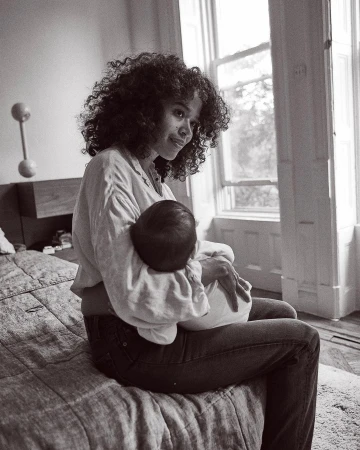 Monochrome portrait of an adult female sitting on a bed, holding a baby in her arms near a window providing natural light.