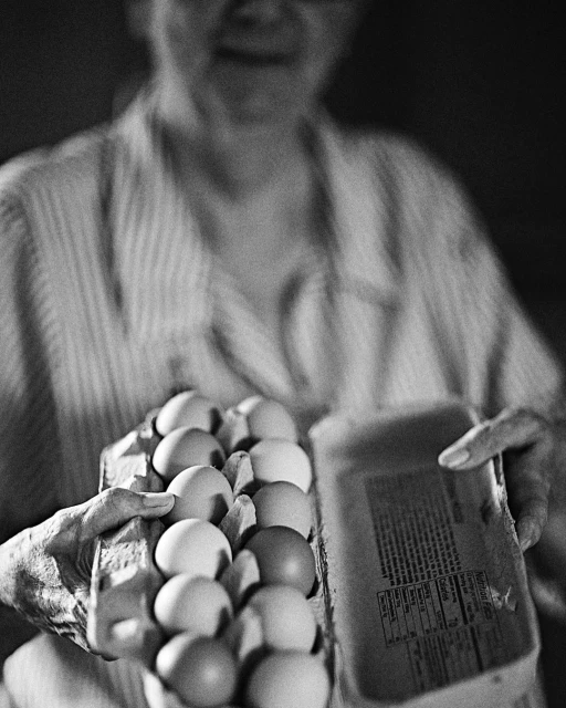 Elderly woman in a striped shirt holds a carton of eggs and a package with nutrition facts label in a black and white photo.