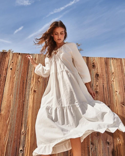 A woman in a white dress stands near a wooden fence under a partly cloudy blue sky, showcasing outdoor fashion with a low-angle perspective.