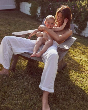 Woman wearing white linen trousers seated outdoors on a wooden chair holding a baby on her lap in Los Angeles.