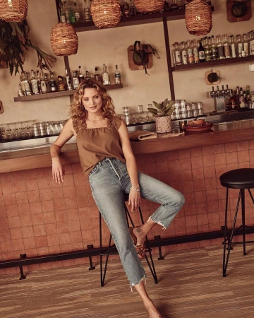 Stylish woman in a brown top and jeans poses in a bar setting with hanging lamps, bottles on shelves, and a tiled counter backdrop.