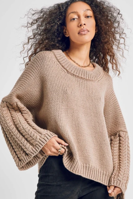 A woman with curly brown hair is wearing a chunky knit sweater with braided sleeve details and a gold chain necklace in a studio shot.