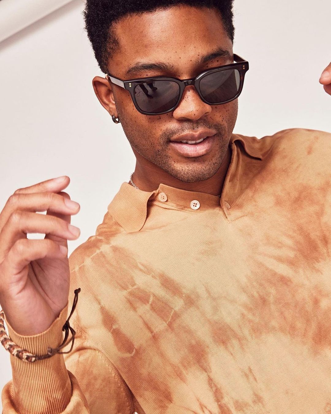 Portrait of a black man wearing dark sunglasses and a tie-dye collared shirt against a light background by Annie Edmonds