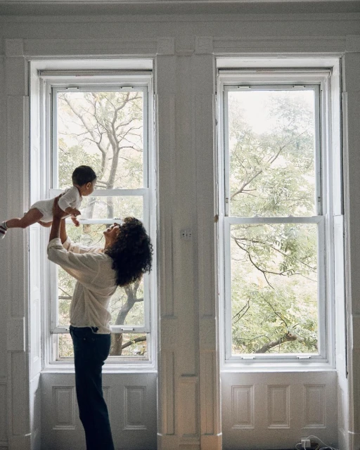A mother with curly hair lifts her infant child up in the air inside a large white room with natural light streaming through large windows.
