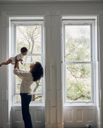 A mother with curly hair lifts her infant child up in the air inside a large white room with natural light streaming through large windows.