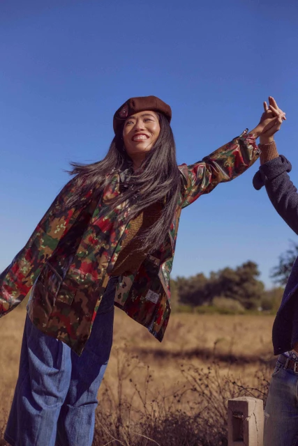 Outdoor shot of two women in a field holding hands under a clear blue sky, one in a patterned jacket and beret, smiling.