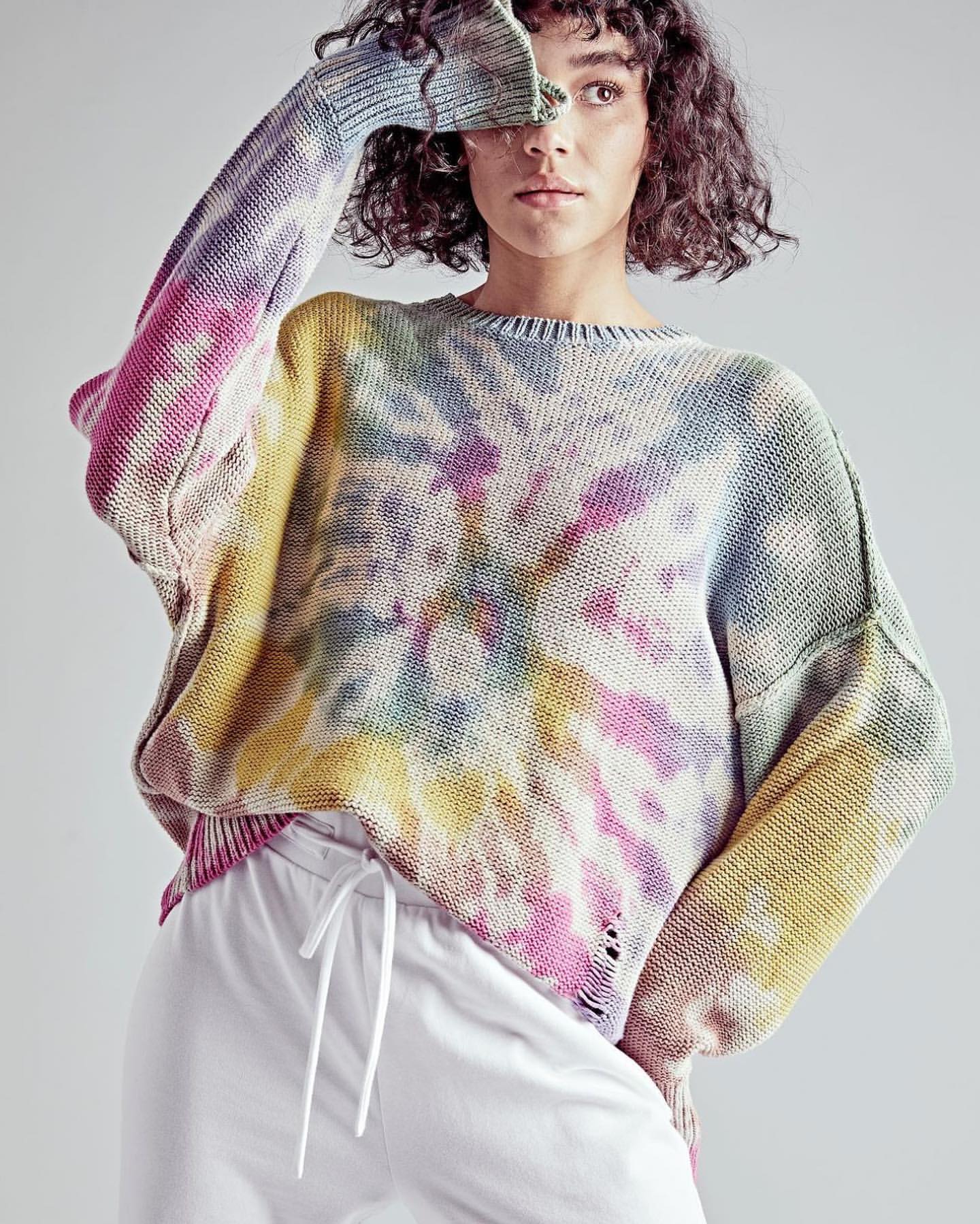 A woman in a tie-dye sweater and white sweatpants poses indoors, obscuring her face with her hand, showcasing a casual fashion style.