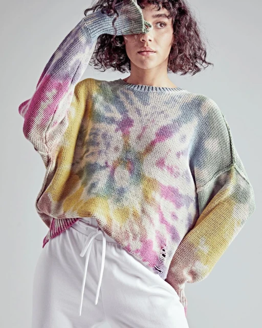 A woman in a tie-dye sweater and white sweatpants poses indoors, obscuring her face with her hand, showcasing a casual fashion style.