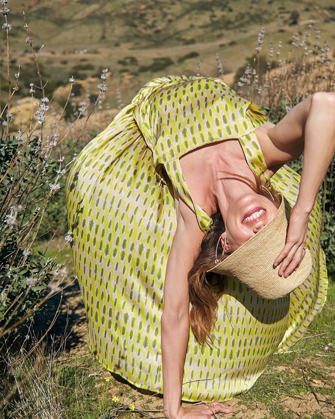 Woman in a yellow patterned dress and straw hat bends backward in a grassy field against a hilly landscape, smiling at the camera.