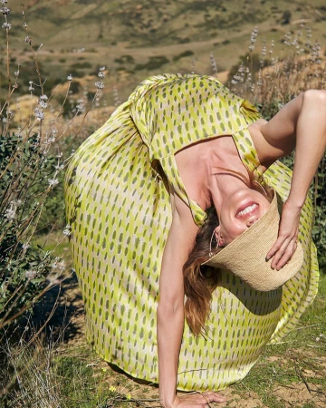 Woman in a yellow patterned dress and straw hat bends backward in a grassy field against a hilly landscape, smiling at the camera.