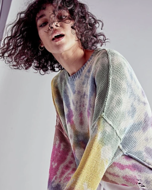 Fashion shot of a curly-haired young woman wearing a tie-dye sweater, looking down at the camera in a neutral grey indoor setting.