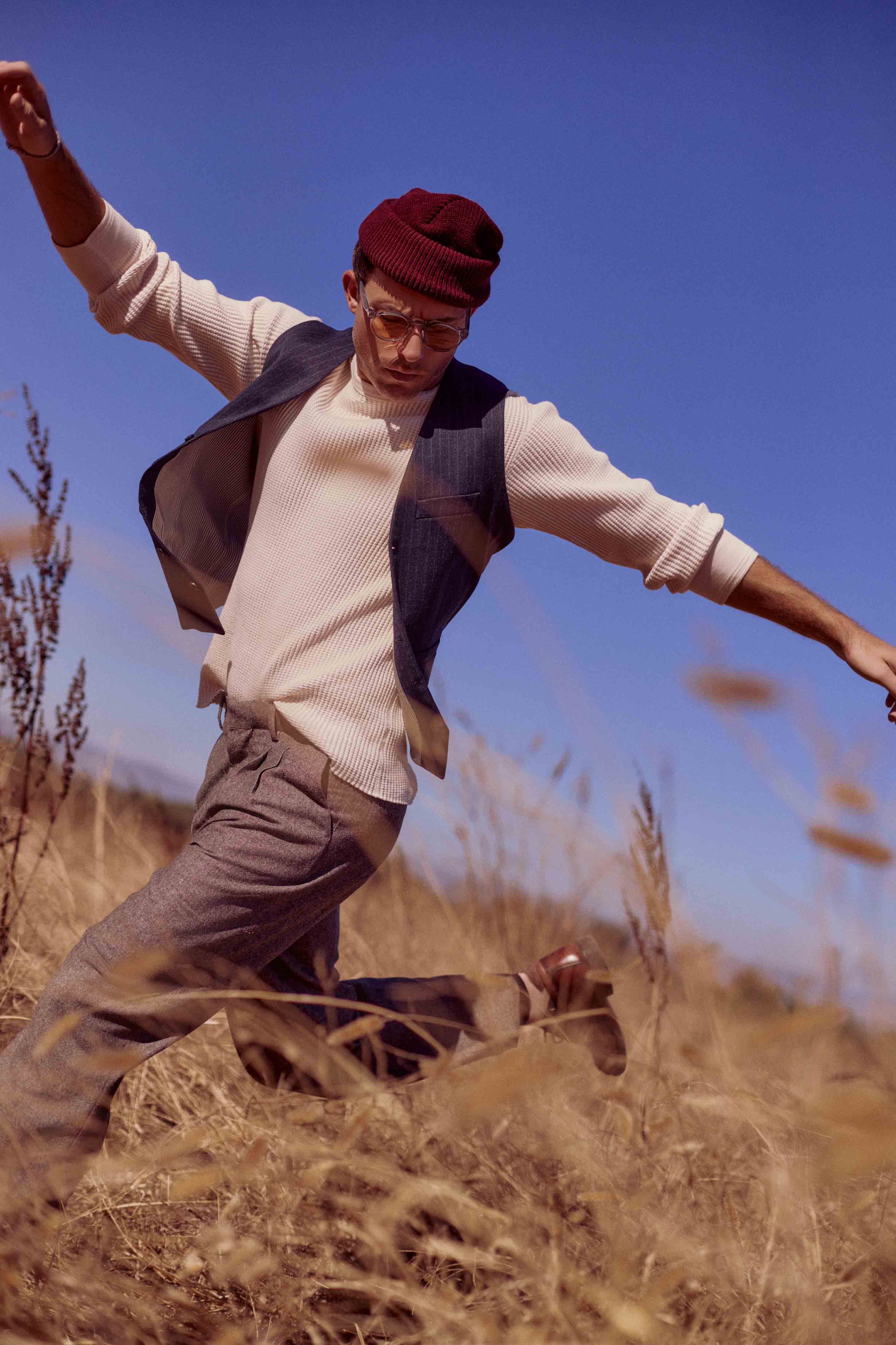 A man wearing a beanie, sweater, and vest jumps in a field of tall grasses under a clear blue sky in an outdoor fashion shot.