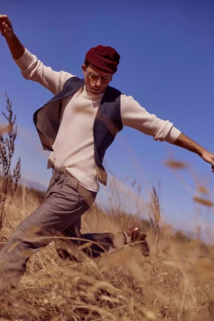 Man wearing a burgundy beanie, white sweater, and vest jumping through dry grass under a blue sky