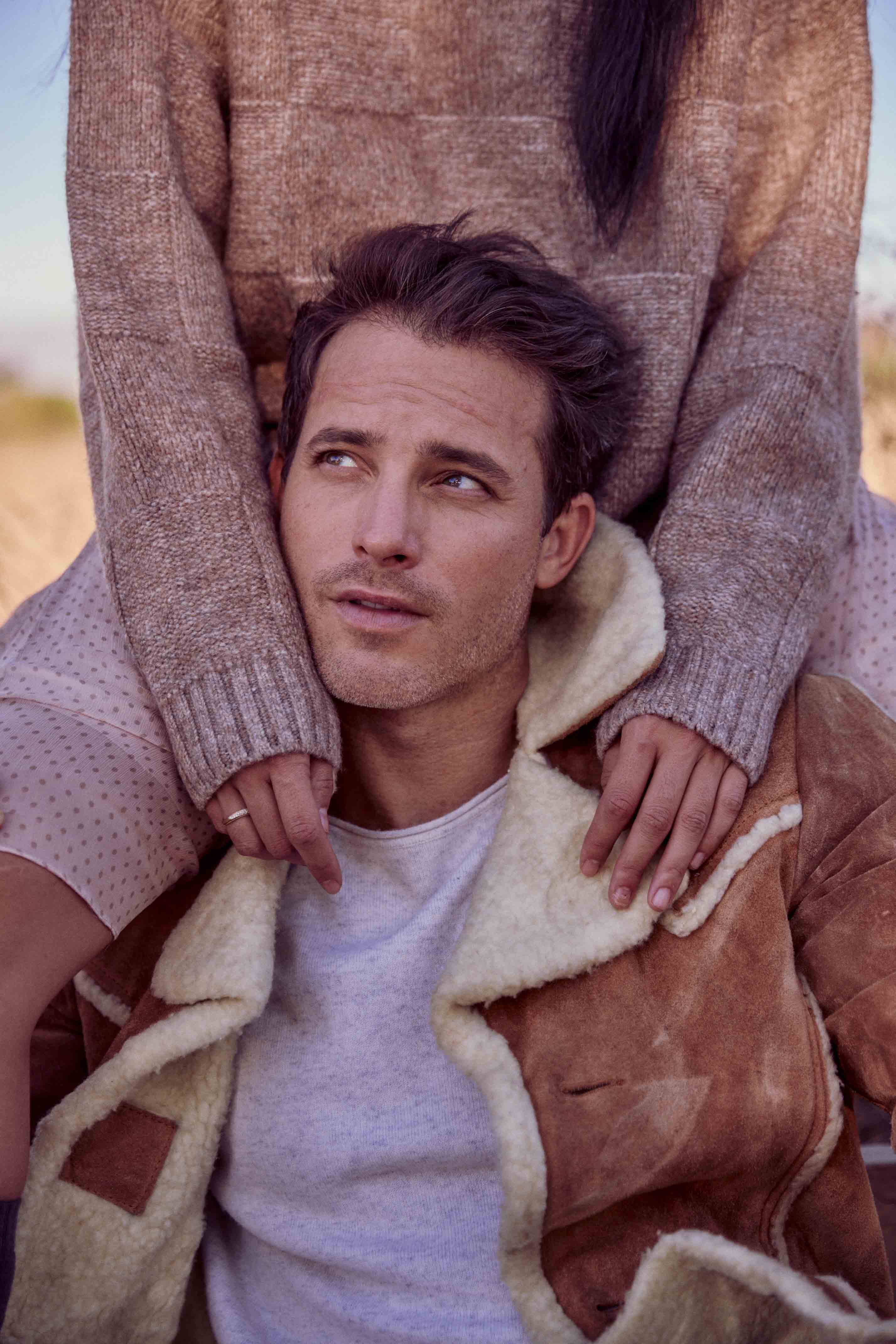 A man wearing a shearling coat with a woman behind him in an outdoor setting. He is looking to the left. The woman is wearing a sweater.